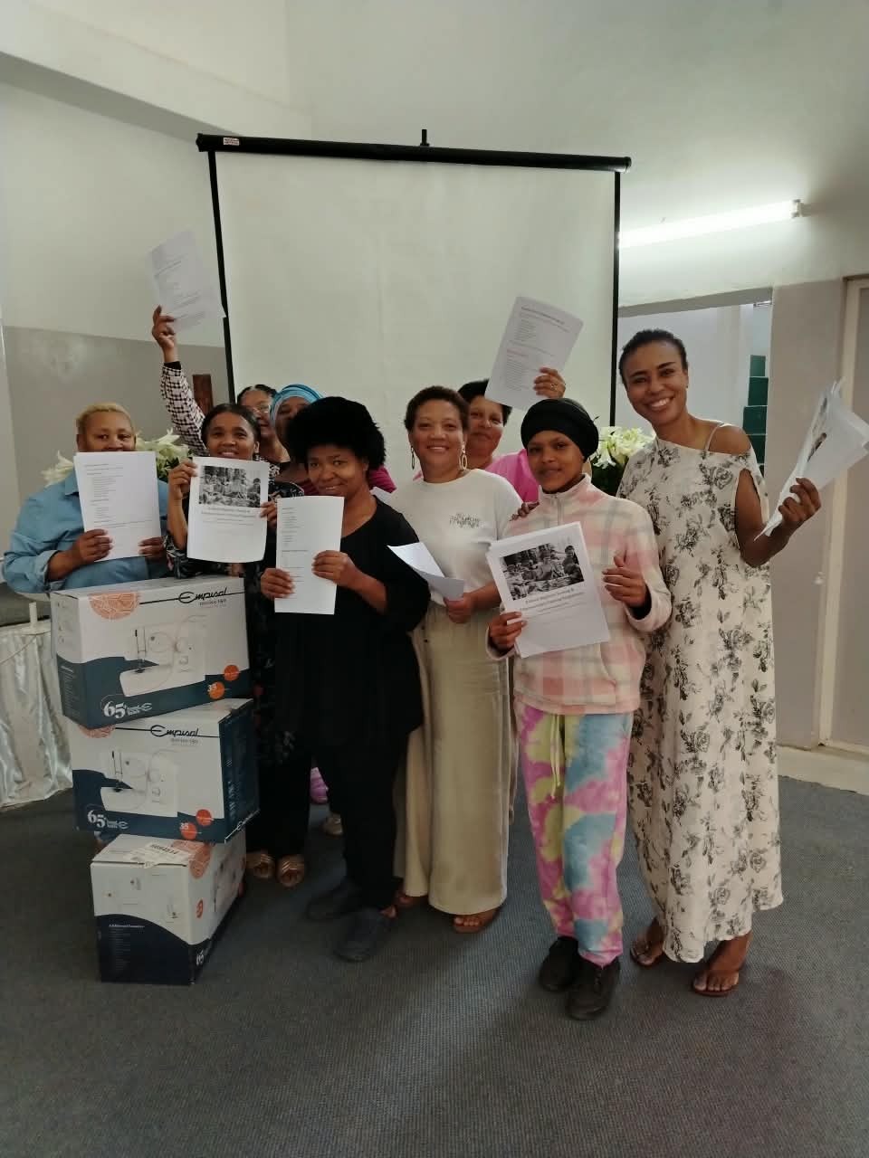 Women in the first sewing class holding their course materials beside new sewing machines